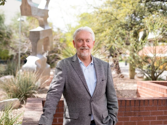 A man in a grey suit poses for a photo in front of desert plants.