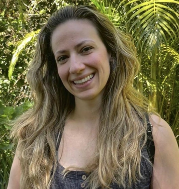 person with long blonde hair wearing a gray tank top, palm foliage in background