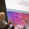 A man in a brown suit stands in front of a podium with his hands in front of him. Behind him is a screen with a presentation that reads "Fusion Research a the University of Arizona"