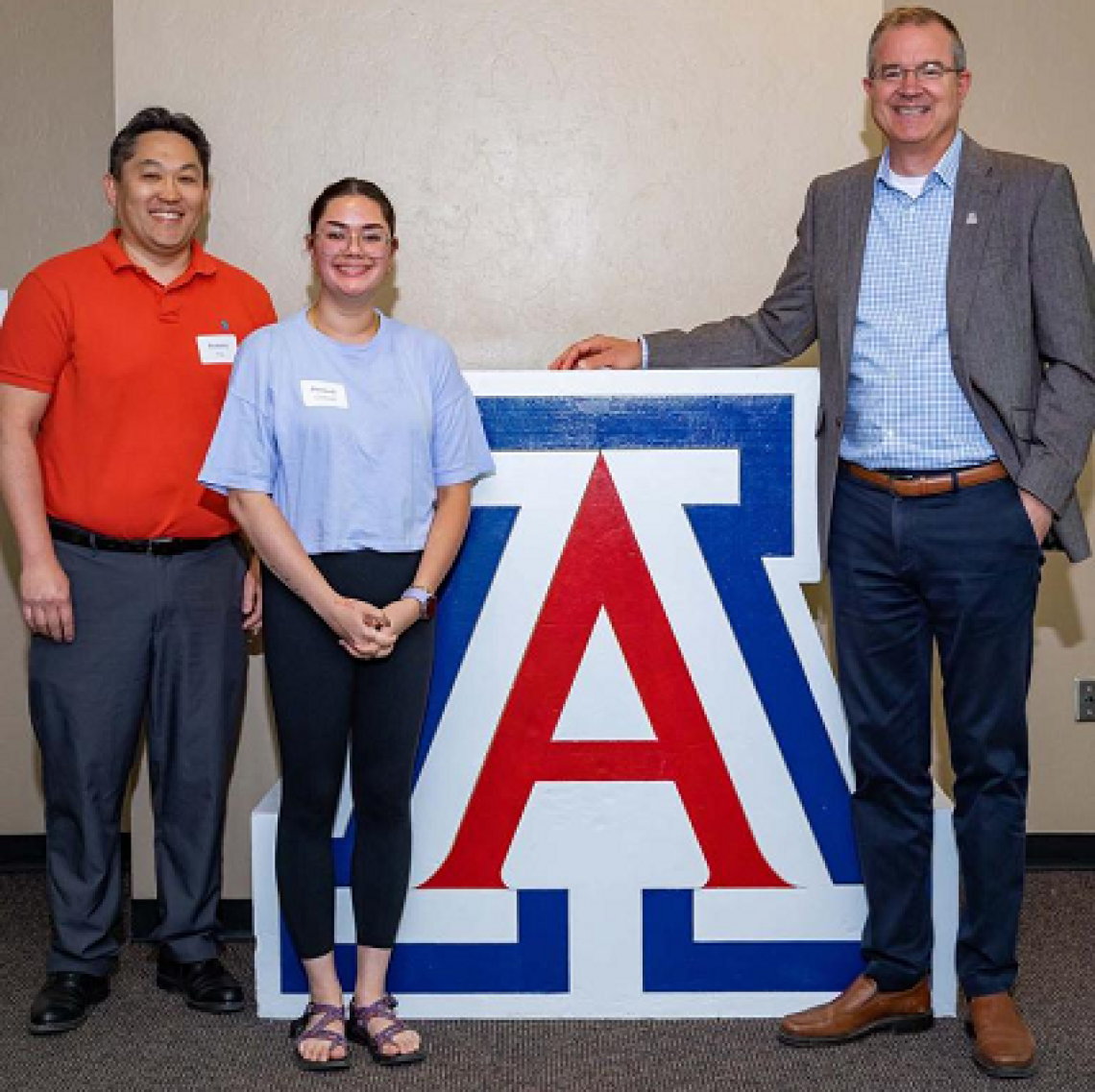 three people, smiling, standing next to UA block A logo