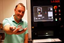 Andrew Wessman stands next to a 3D printer and holds out a 3D-printed metal part.
