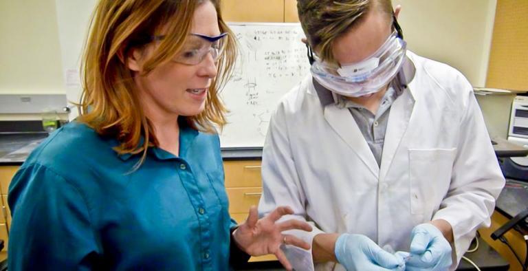 A professor stands next to a student in a lab coat, explaining something. Both are wearing safety goggles.